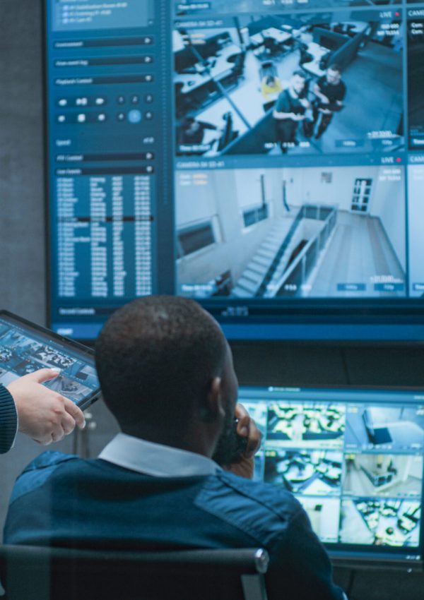 Female security worker talks with African American colleague, uses tablet. Multiethnic CCTV operators control security cameras on computer monitors and big digital screen with surveillance footage.